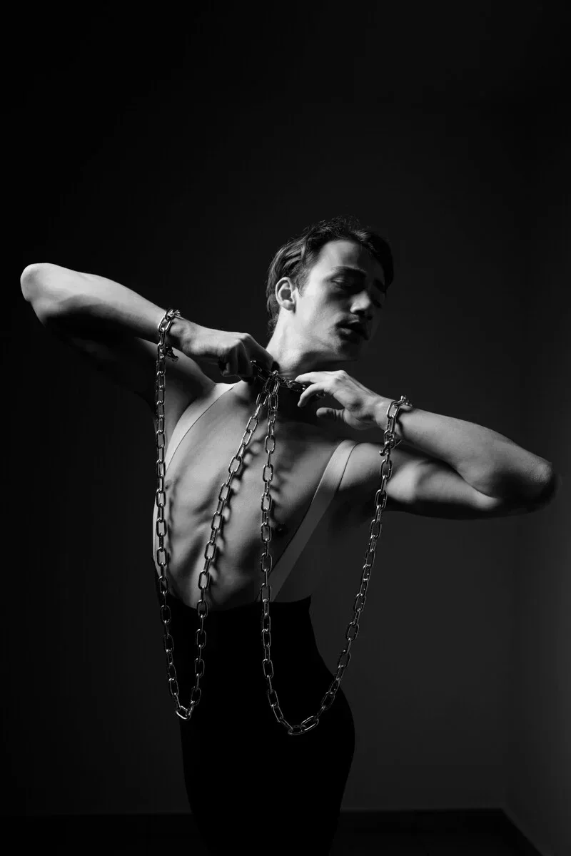 Uno studio monocromatico ad alto contrasto di un artista uomo in una posa ballettistica di resistenza, a simboleggiare i paesaggi psicologici della condizione umana e la liberazione creativa.