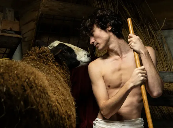 Profile of a shirtless young man holding a wooden staff and looking at a brown sheep in a dark stable.