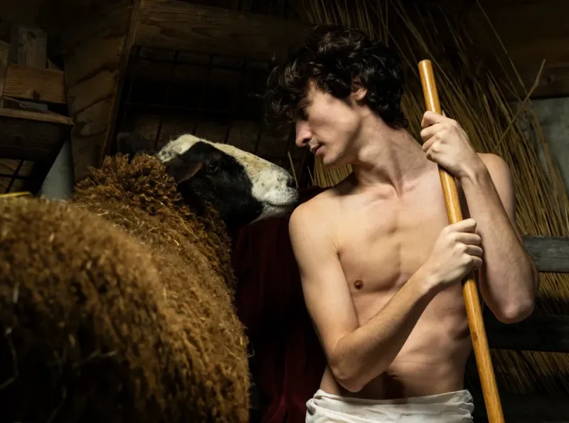 Profile of a shirtless young man holding a wooden staff and looking at a brown sheep in a dark stable.
