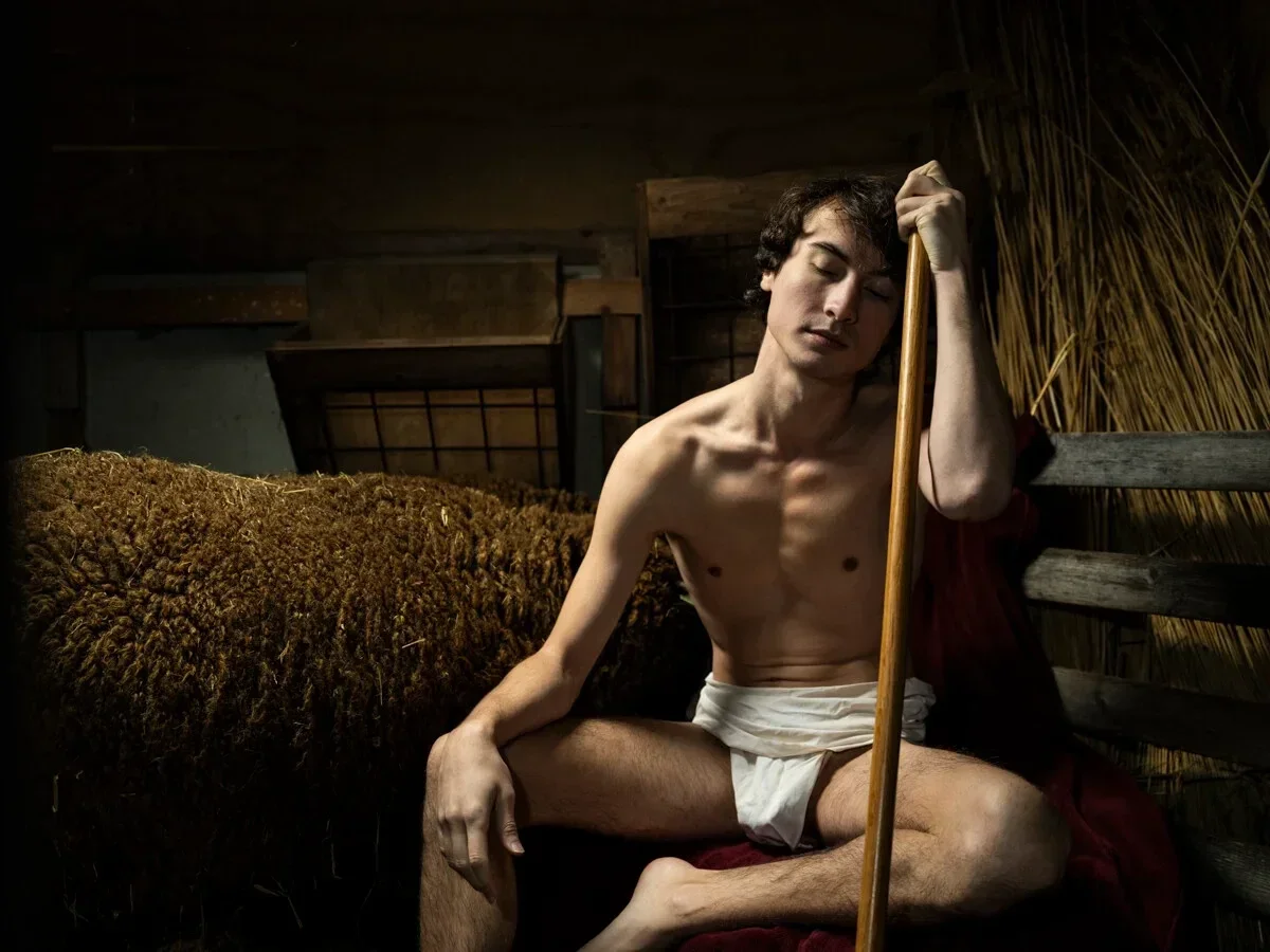 Jeune homme aux yeux fermés, adossé à un bâton en bois dans une étable sombre, à côté d'un mouton.