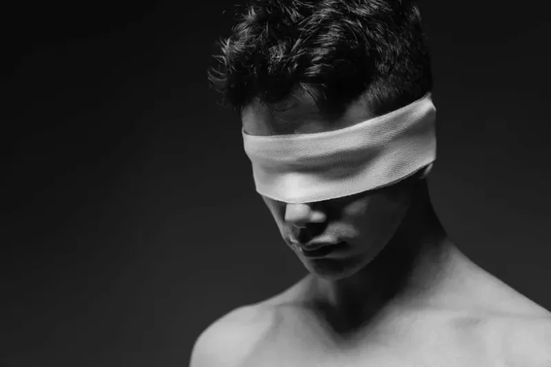 A minimalist black and white conceptual portrait of a man wearing a blindfold, emphasizing shadows and skin texture.