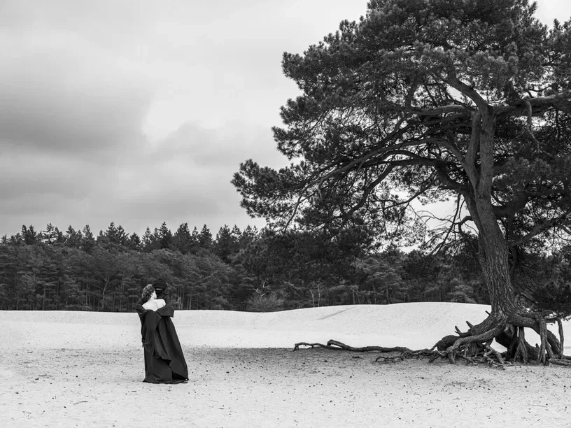 Una fotografía de bellas artes en blanco y negro de Arjan Spannenburg titulada ROOTED, que presenta a un hombre de hombros descubiertos y una figura con túnicas oscuras de Oriente Medio abrazándose en una vasta duna de arena junto a un pino escocés erosionado con raíces expuestas.