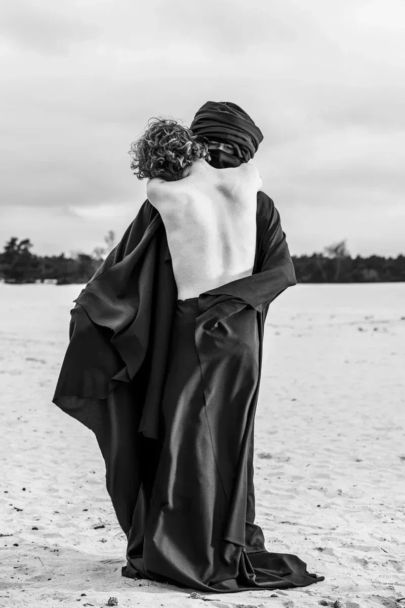 A close embrace between a bare-chested man and a figure in dark traditional robes on a bright sandy beach.