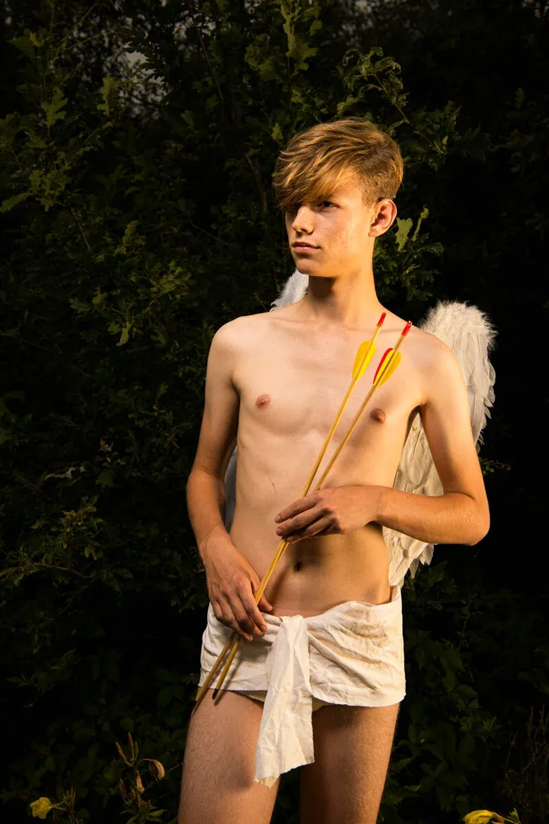 Fine art portrait of a young male model dressed as Cupid holding arrows, standing in a dark forest setting, representing the piece 'The Question of the Heart'.
