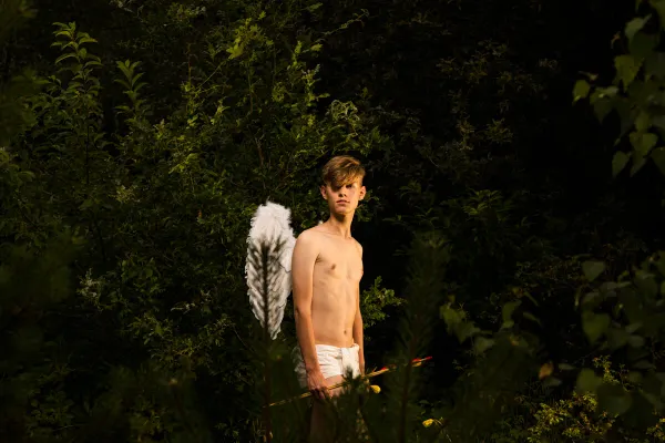 A young man with wings and a bow stands among dark leaves, embodying a vulnerable contemporary Cupid.