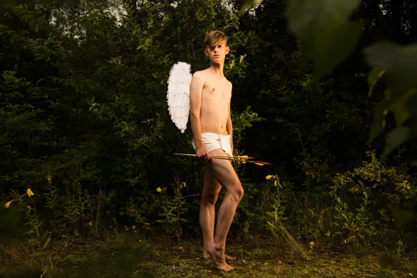 Fine art color photograph titled 'The Quest' featuring a young man with white wings and arrows posing as Cupid in a dark forest.