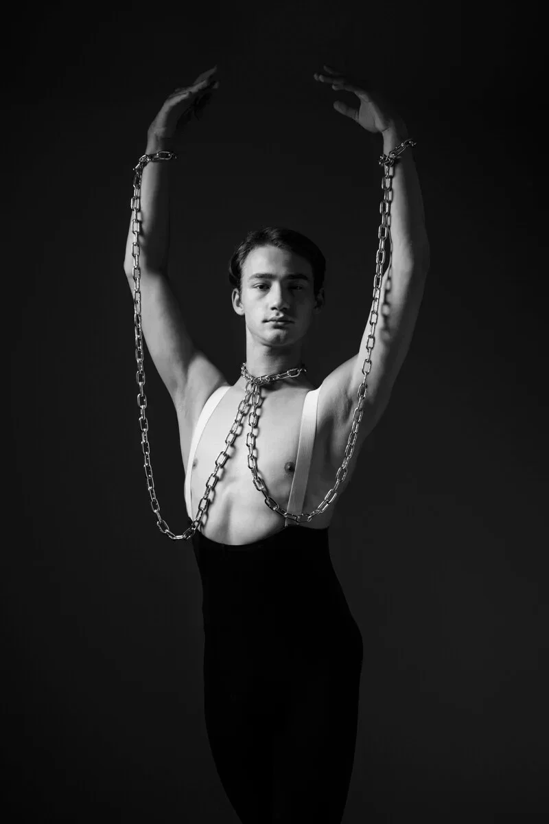 Un ritratto verticale in bianco e nero di ARJAN SPANNENBURG, intitolato BOUND II, che mostra un uomo di fronte alla macchina da presa con uno sguardo diretto e pesanti catene intorno al collo e ai polsi.