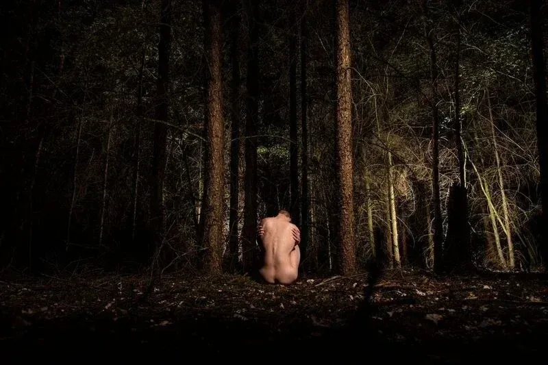 A nude figure in a protective fetal position at night, illuminated by a spotlight in a dark forest; a photographic metaphor for being overwhelmed by emotion.
