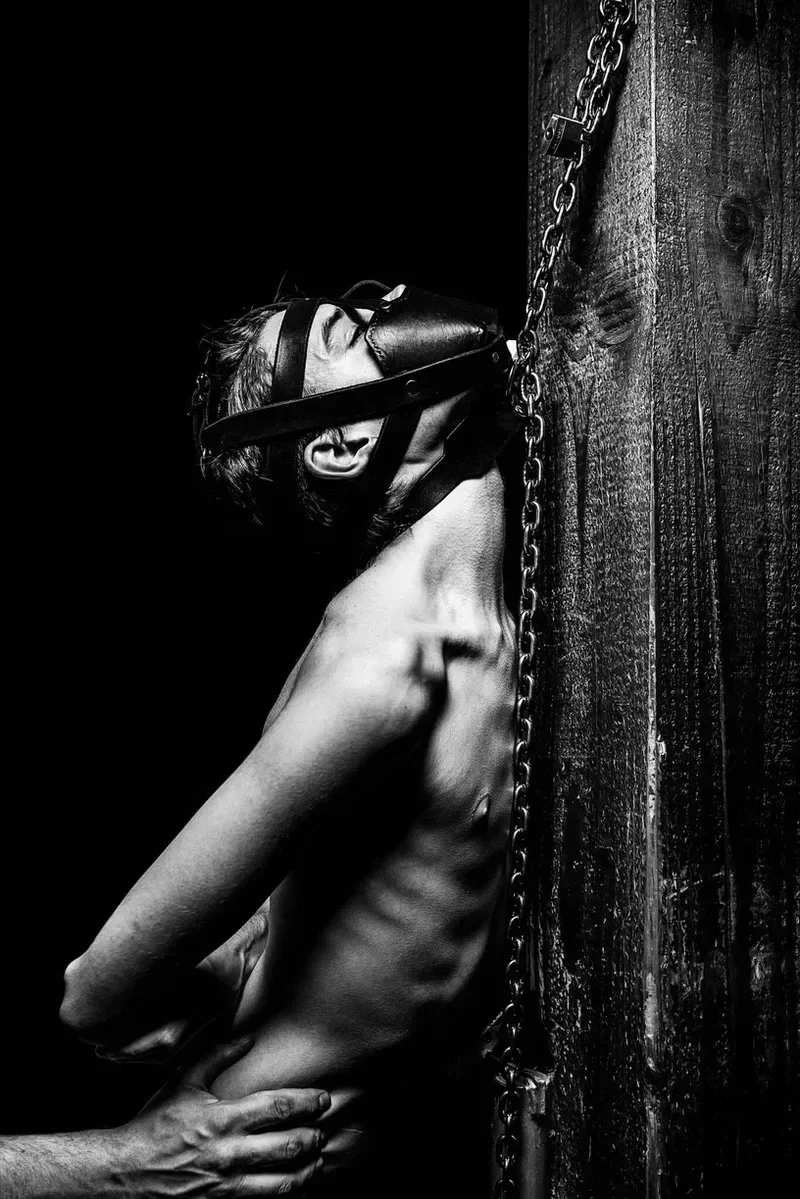 Arjan Spannenburg shows a dramatic black and white fine art photograph titled "LOVED," featuring a lean figure in a state of intense surrender. The subject wears a leather mask and is connected to a rustic wooden post by a heavy metal chain. Below, a pair of hands gently yet firmly cups the subject's waist, providing a grounding, protective touch that contrasts with the themes of restraint and vulnerability.