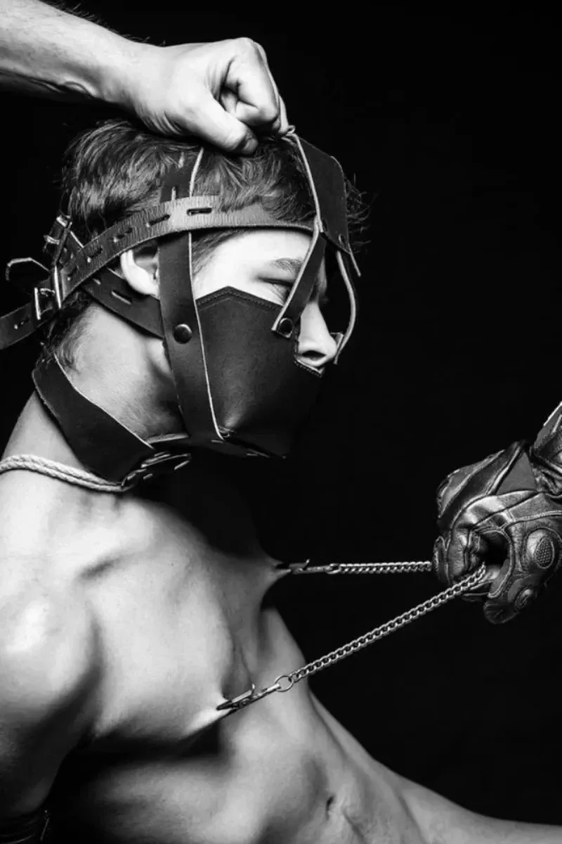 Arjan Spannenburg's "PERSISTANCE": B/W image of a masked man with chains, hand adjusting headgear. Provocative art.