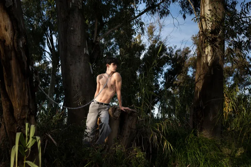 A conceptual fine art portrait of a shirtless young man chained to a tree in a sun-dappled forest, symbolizing the struggle to hold onto youth and the transience of beauty.