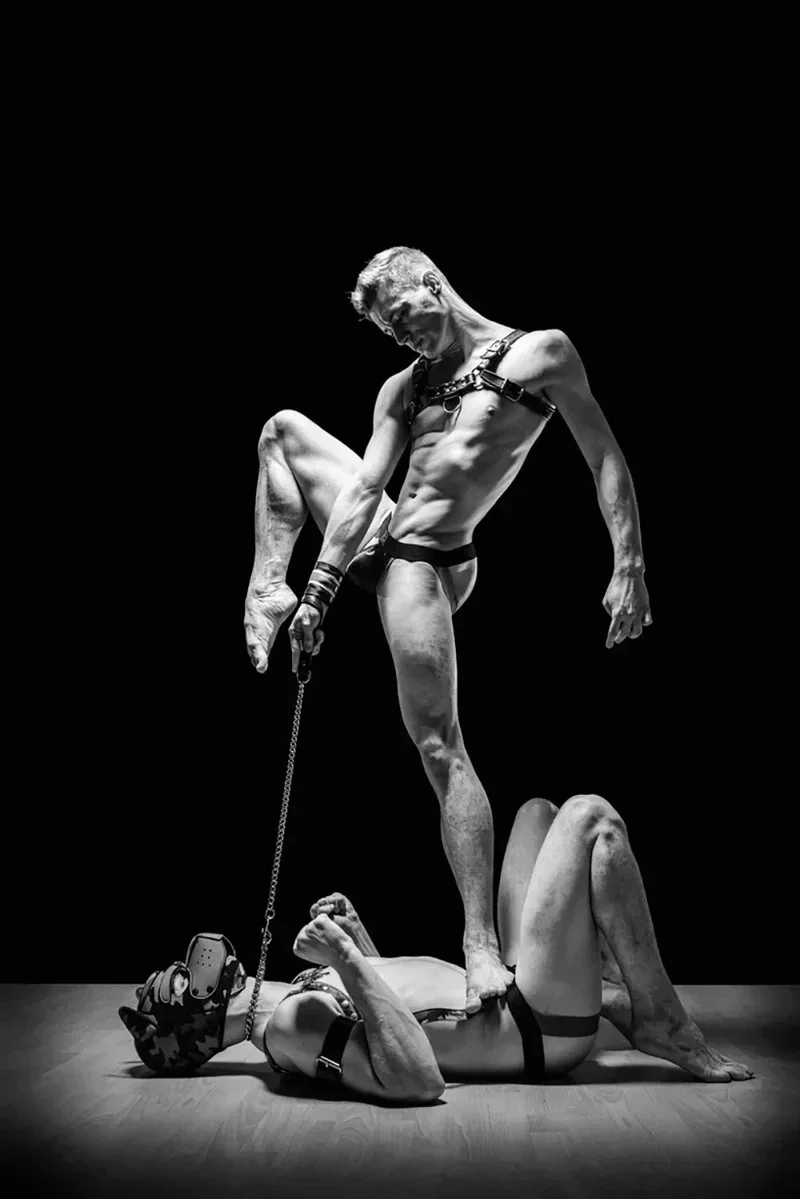 Et sort-hvidt Fine Art Print med titlen RECIPROCATION af Arjan Spannenburg. Billedet viser en højt koreograferet, skulpturel interaktion mellem to mænd på en mørk scene. Den ene mand står i en dynamisk, atletisk positur, balanceret med den ene fod på brystet af en mand, der ligger under ham. Den stående figur bærer en lædersele og holder en kæde, der er forbundet til kraven på den nederste figur, som bærer en camouflagehætte. Den høj-kontrast belysning fremhæver muskeldefinitionen og den bevidste, gensidige tillid i udførelsen.