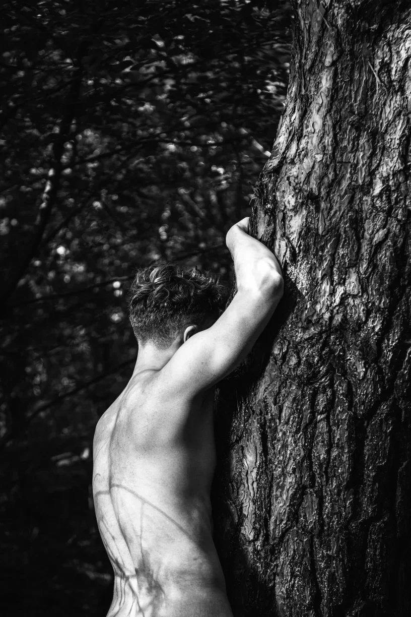 Una fotografía de bellas artes en blanco y negro que captura una espalda masculina apoyada en el tronco de un árbol texturizado, enfatizando la textura de la piel y las sombras del follaje para representar la vulnerabilidad masculina y una conexión con la naturaleza.