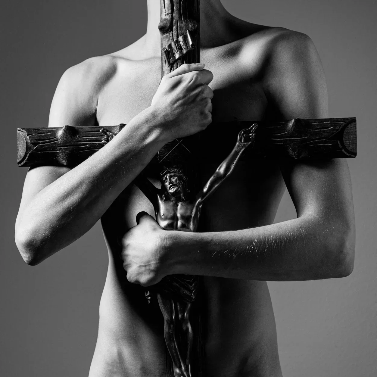 Das Kunstwerk SEGREGATION; eine kontrastreiche Schwarz-Weiß-Fotografie in Museumsqualität, die eine Person zeigt, die ein hölzernes Kruzifix vor ihre nackte Brust hält.
